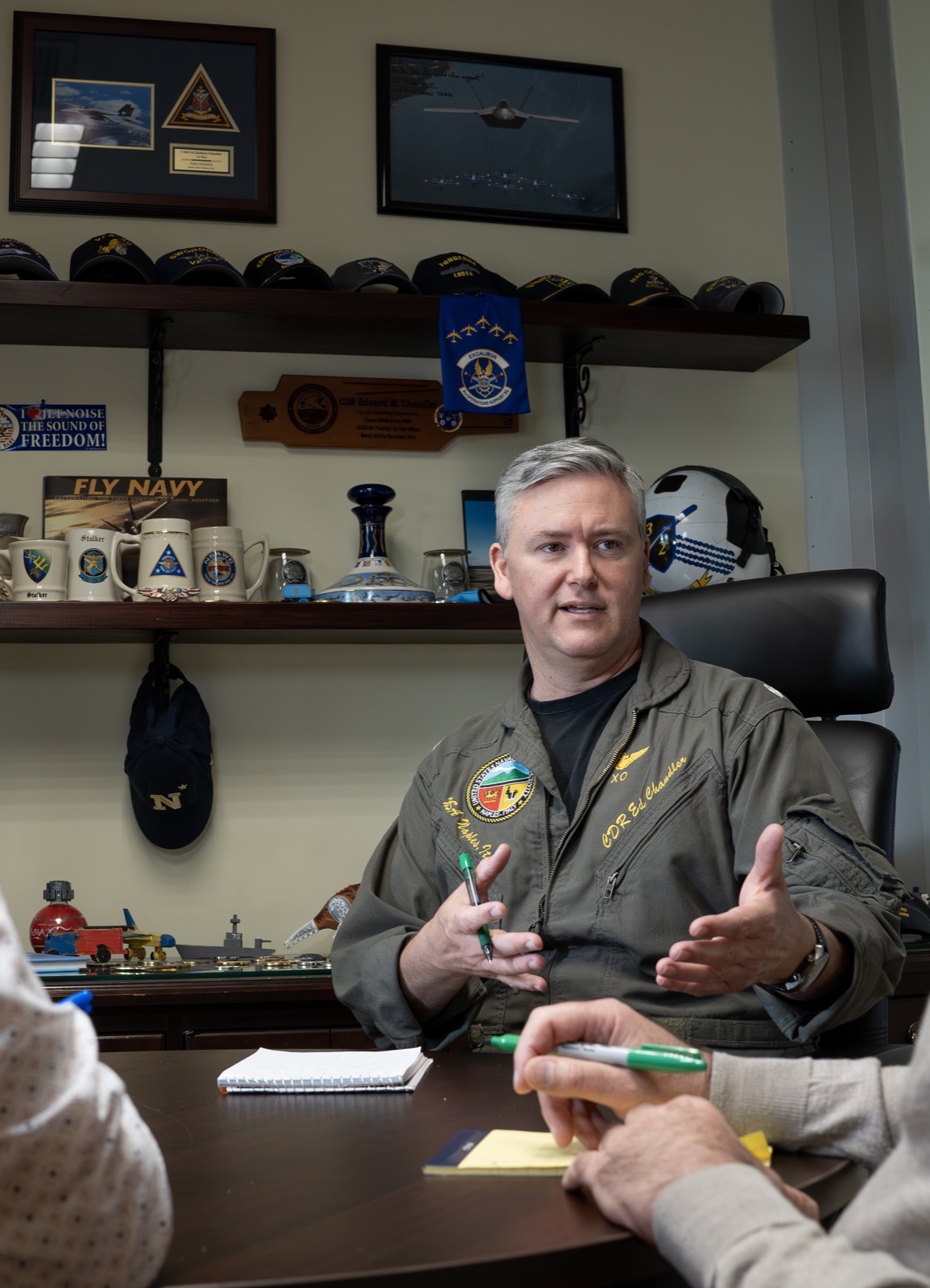 Ed Chandler mid-conversation in his office, two colleagues at the table and mementos from his Navy career behind him